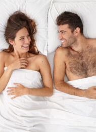 Happy newlywed couple enjoy comfort in bed, look at each other with love and joy, lying in bed under white bedclothes, make plans for future, have romantic relationships, feel good and relaxed