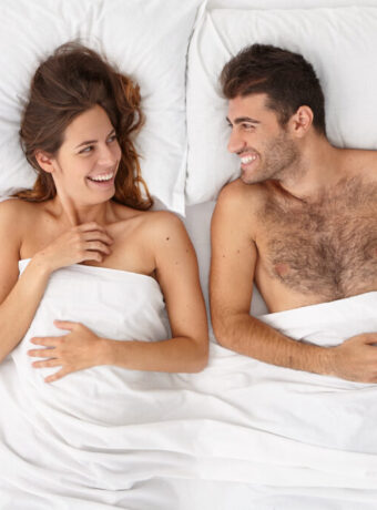 Happy newlywed couple enjoy comfort in bed, look at each other with love and joy, lying in bed under white bedclothes, make plans for future, have romantic relationships, feel good and relaxed